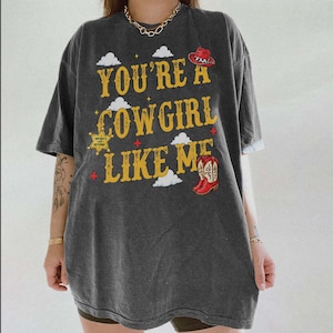 May include: A charcoal gray oversized t-shirt featuring the yellow text "YOU'RE A COWGIRL LIKE ME" with graphics of a red cowboy hat, a sheriff's star badge with "INFINITY AND BEYOND" text, red cowboy boots, and white clouds on the front.