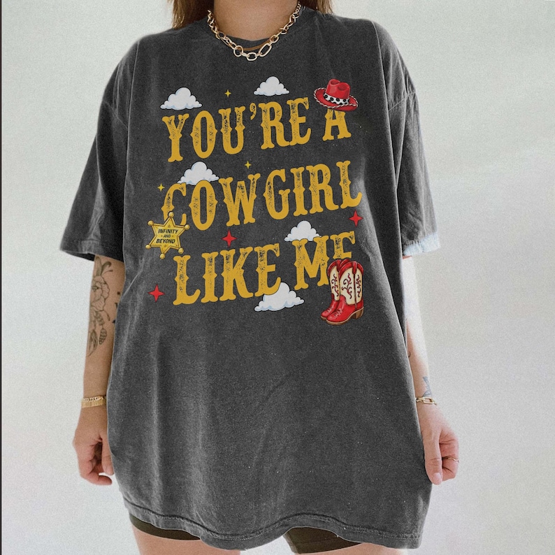 May include: A charcoal gray oversized t-shirt featuring the yellow text "YOU'RE A COWGIRL LIKE ME" with graphics of a red cowboy hat, a sheriff's star badge with "INFINITY AND BEYOND" text, red cowboy boots, and white clouds on the front.