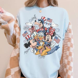 May include: Pale blue sweatshirt with a patriotic Disney design. It features Mickey Mouse, Minnie Mouse, Donald Duck, Goofy, and Pluto, along with American flags, fireworks, and text commemorating the USA's 250th anniversary.
