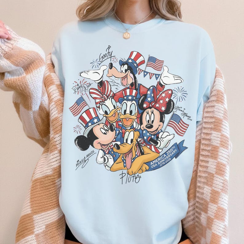 May include: Pale blue sweatshirt with a patriotic Disney design. It features Mickey Mouse, Minnie Mouse, Donald Duck, Goofy, and Pluto, along with American flags, fireworks, and text commemorating the USA's 250th anniversary.