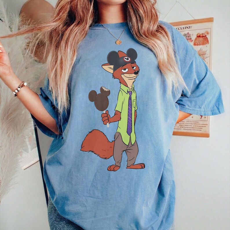 Puede incluir: Camiseta azul claro con un personaje de zorro de dibujos animados que lleva un sombrero con orejas de rat&oacute;n y sostiene un helado de chocolate. El zorro viste una camisa verde, una corbata morada y pantalones grises. La camiseta es holgada.