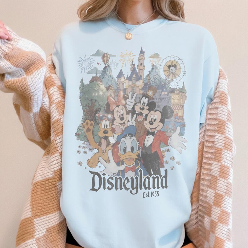 Puede incluir: Sudadera azul claro con cuello redondo y un gr&aacute;fico de Disneyland con Mickey Mouse, Minnie Mouse, Donald Duck y Pluto. El gr&aacute;fico incluye un castillo, fuegos artificiales y el texto "Disneyland Est. 1955".