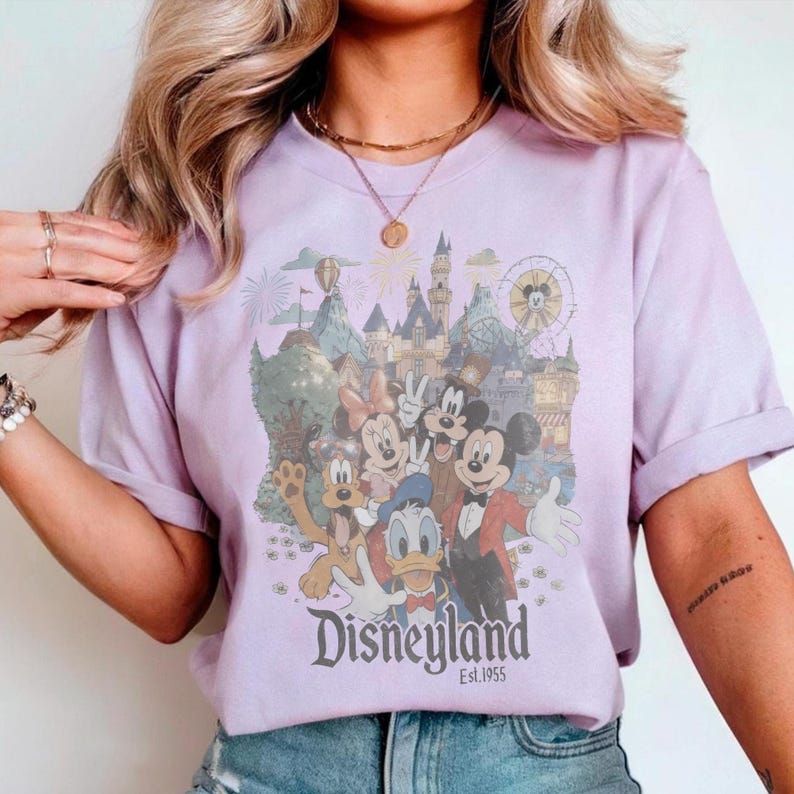 Puede incluir: Una camiseta de color lila con un dise&ntilde;o de Disneyland que presenta a Mickey Mouse, Minnie Mouse, Donald Duck y Goofy. El dise&ntilde;o incluye un castillo, fuegos artificiales y una noria. El texto "Disneyland Est. 1955" est&aacute; impreso debajo.