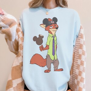 Puede incluir: Sudadera azul claro con un personaje de zorro de dibujos animados que lleva un sombrero de Mickey Mouse y sostiene un helado con forma de Mickey Mouse. El zorro viste una camisa verde, una corbata morada y pantalones marrones.