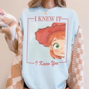 以下が含まれることがあります： 赤髪で赤いカウボーイハットをかぶった漫画のキャラクターのグラフィックプリントが施された、ライトブルーのクルーネックトレーナー。グラフィックの上には「I KNEW IT」、下には「I Knew You」という文字が赤い長方形の枠内にプリントされています。