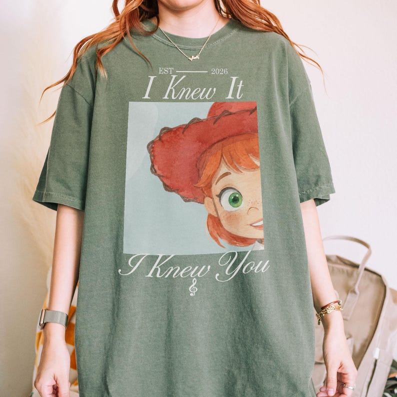 K&ouml;nnte beinhalten: Ein olivgr&uuml;nes Kurzarm-T-Shirt mit einem Grafikdruck einer Zeichentrickfigur mit roten Haaren und einem roten Cowboyhut. Der Text lautet "EST 2026", "I Knew It" und "I Knew You", darunter befindet sich ein kleines Musiknotensymbol.