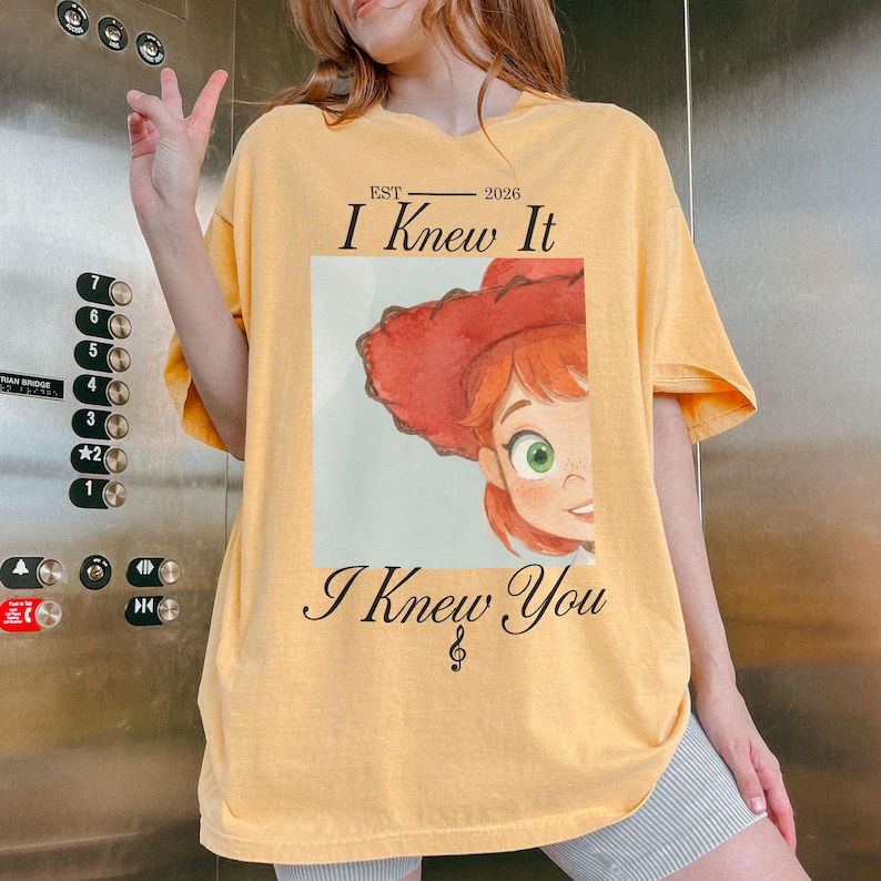 K&ouml;nnte beinhalten: Ein hellgelbes T-Shirt mit kurzen &Auml;rmeln und einem Grafikdruck einer Zeichentrickfigur mit roten Haaren und einem roten Hut. Der Text EST 2026 I Knew It und I Knew You mit einer Musiknote ist in Schwarz &uuml;ber und unter der Grafik aufgedruckt.