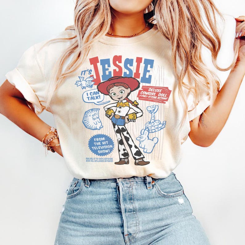 May include: Cream-colored t-shirt featuring a graphic of the character Jessie from the animated film "Toy Story." The design includes the name "Jessie" in red and blue, along with cartoon illustrations and the text "I CAN TALK!" and "DELUXE COWGIRL DOLL."