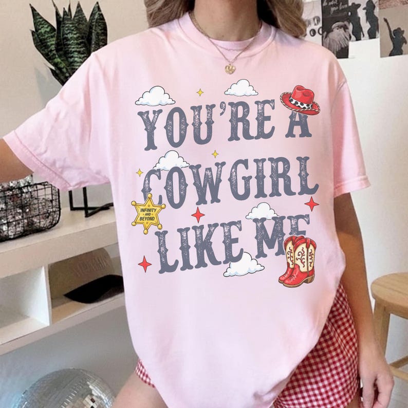 May include: A light pink t-shirt featuring the text "YOU'RE A COWGIRL LIKE ME" in dark gray lettering. The design includes white clouds, red stars, a yellow sheriff badge with "INFINITY AND BEYOND" text, a red cowboy hat, and red cowboy boots.