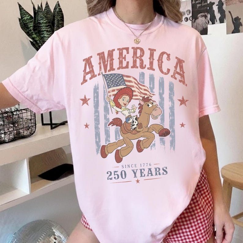 May include: A light pink t-shirt featuring a graphic print of a cowgirl riding a horse while holding an American flag. The text reads AMERICA, SINCE 1776, and 250 YEARS. The design includes red and blue stars and a faded vertical striped background.