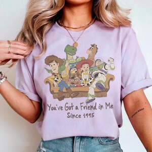 May include: A light purple t-shirt featuring a graphic of Toy Story characters sitting on a couch. The text reads, You've Got a Friend in Me Since 1995. The characters include Woody, Buzz Lightyear, Jessie, Rex, Slinky Dog, Mr. Potato Head, and Bullseye.