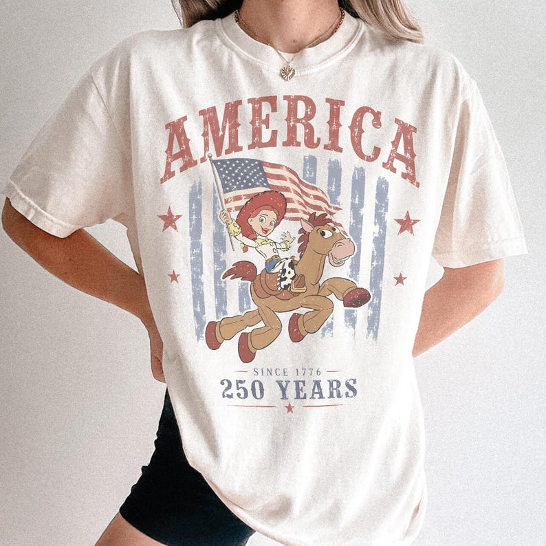 May include: A cream-colored short-sleeve t-shirt featuring a graphic print of a cowgirl riding a brown horse while holding an American flag. The text reads AMERICA, SINCE 1776, and 250 YEARS, accompanied by red and blue star and stripe accents.