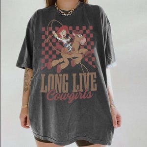 K&ouml;nnte beinhalten: Dunkelgraues T-Shirt mit einem Cartoon-Bild einer Cowgirl, die auf einem Pferd reitet und eine Lasso schwingt, vor einem rot-braunen Schachbrettmuster. Der Text "LONG LIVE Cowgirls" ist darunter gedruckt.