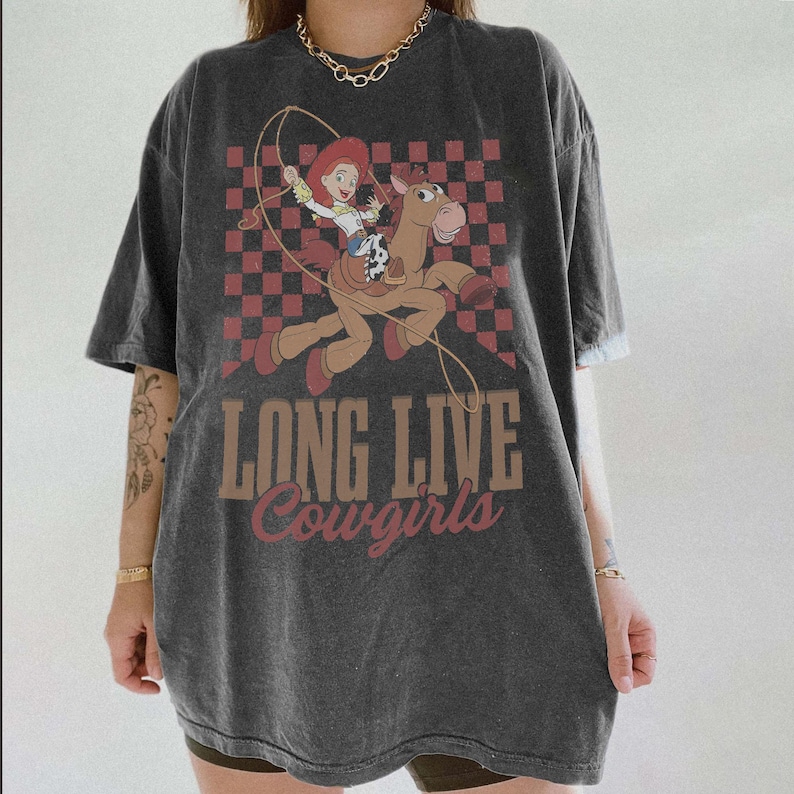 K&ouml;nnte beinhalten: Dunkelgraues T-Shirt mit einem Cartoon-Bild einer Cowgirl, die auf einem Pferd reitet und eine Lasso schwingt, vor einem rot-braunen Schachbrettmuster. Der Text "LONG LIVE Cowgirls" ist darunter gedruckt.