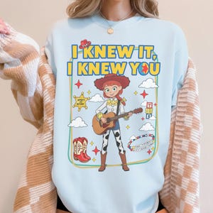 May include: A light blue crewneck sweatshirt featuring a graphic of a cowgirl character playing an acoustic guitar. The text reads I KNEW IT, I KNEW YOU. Surrounding elements include a cowboy hat, a sheriff star, alphabet blocks, boots, and a beaded bracelet.