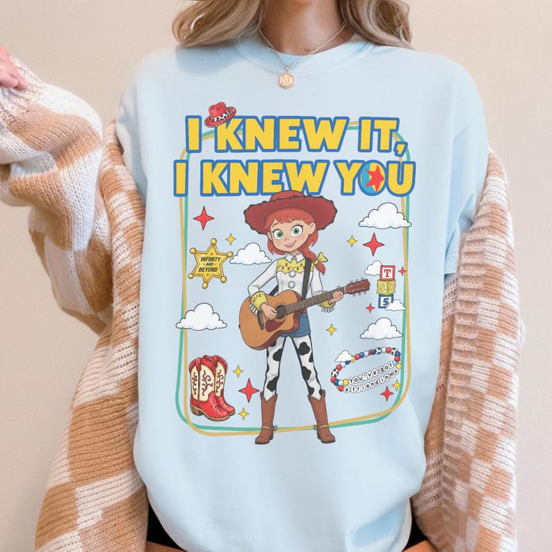 May include: A light blue crewneck sweatshirt featuring a graphic of a cowgirl character playing an acoustic guitar. The text reads I KNEW IT, I KNEW YOU. Surrounding elements include a cowboy hat, a sheriff star, alphabet blocks, boots, and a beaded bracelet.