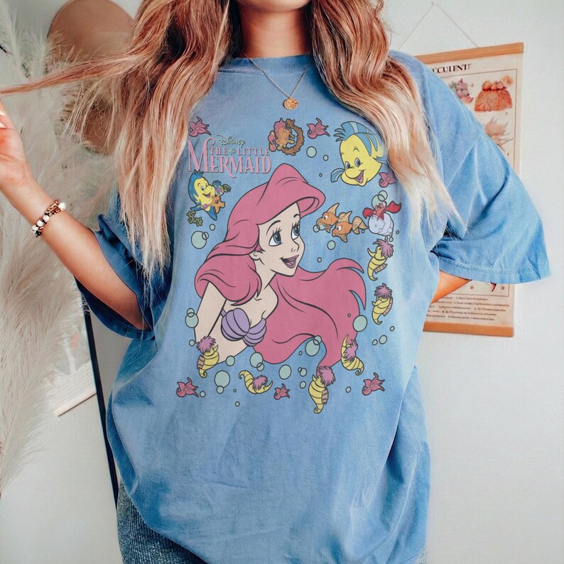 May include: Light blue t-shirt featuring a graphic of Ariel from Disney's The Little Mermaid. The design includes Ariel with pink hair, surrounded by sea creatures. The shirt has short sleeves and a relaxed fit.