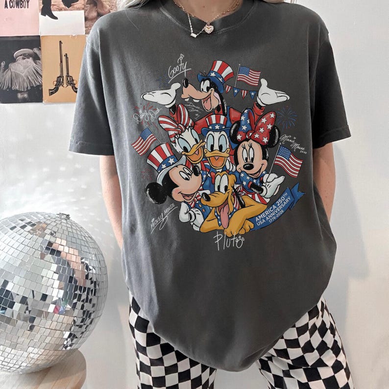May include: Dark grey t-shirt with a graphic of Disney characters Mickey Mouse, Minnie Mouse, Donald Duck, Daisy Duck, Goofy, and Pluto, with American flags and text. The shirt commemorates the USA's 250th anniversary.