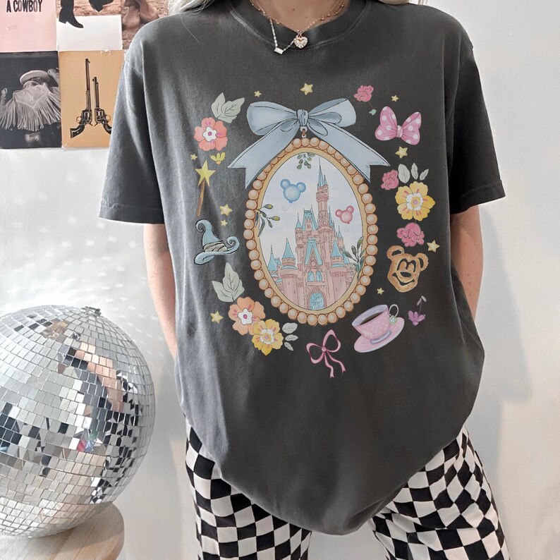 May include: Dark grey t-shirt with a colourful illustration of a castle in an oval frame, surrounded by flowers, bows, and character icons. The design includes a light blue bow and a pink teacup. The shirt has short sleeves.