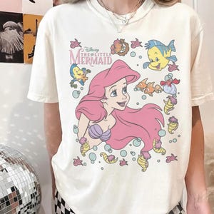 May include: Cream-colored t-shirt featuring a graphic of Ariel from Disney's The Little Mermaid. Ariel has pink hair and a purple seashell bra. The design includes cartoon fish and bubbles. The text "Disney The Little Mermaid" is above Ariel.
