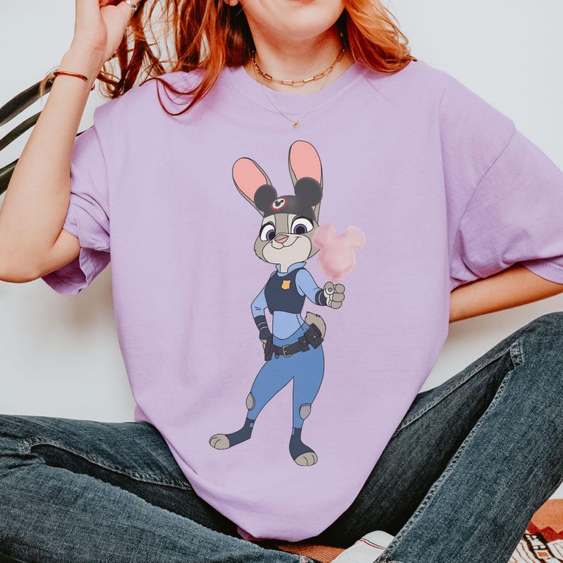 Puede incluir: Camiseta de color lila claro con una imagen de dibujos animados de Judy Hopps de Zootopia. Judy lleva un uniforme de polic&iacute;a y sostiene un algod&oacute;n de az&uacute;car rosa. La camiseta es de estilo casual.