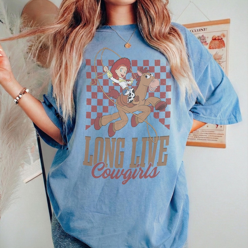 K&ouml;nnte beinhalten: Hellblaues T-Shirt mit einem Cartoon-Motiv einer Cowgirl auf einem Pferd vor einem rot-blauen Karomuster. Unter dem Motiv steht der Text "LONG LIVE Cowgirls" in brauner Schrift.