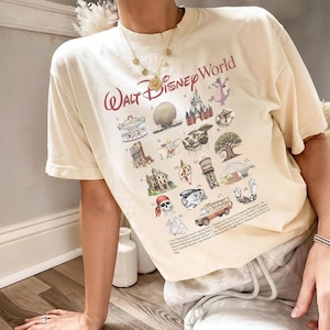 K&ouml;nnte beinhalten: Cremefarbenes T-Shirt mit dem Schriftzug "Walt Disney World" in burgunderroter Schrift. Das Shirt zeigt verschiedene Disney-Park-Symbole und -Attraktionen in einem farbenfrohen, illustrierten Stil, darunter ein Karussell, Spaceship Earth und ein Bus.