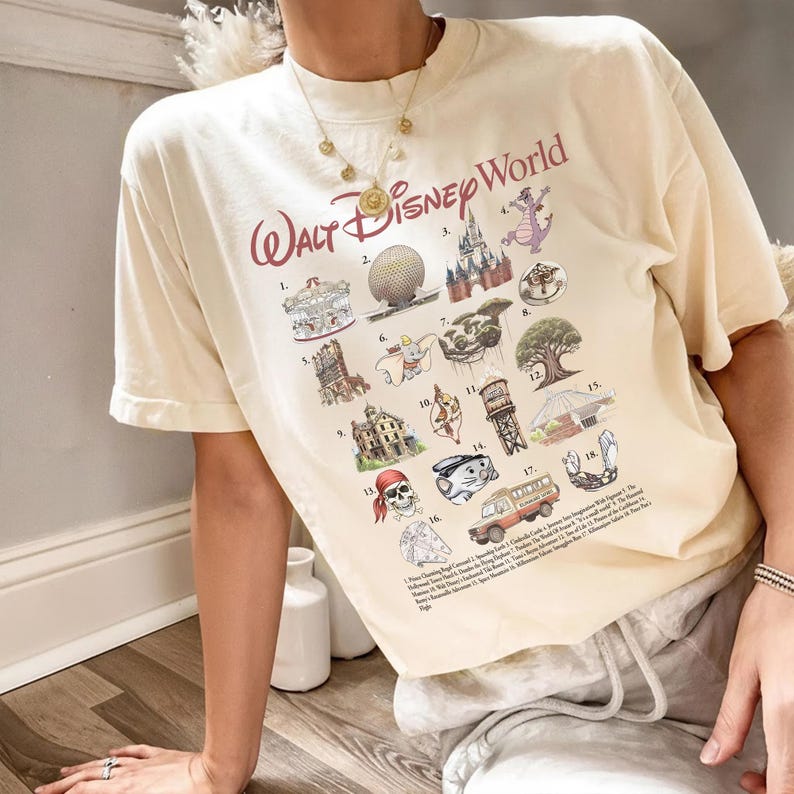 K&ouml;nnte beinhalten: Cremefarbenes T-Shirt mit dem Schriftzug "Walt Disney World" in burgunderroter Schrift. Das Shirt zeigt verschiedene Disney-Park-Symbole und -Attraktionen in einem farbenfrohen, illustrierten Stil, darunter ein Karussell, Spaceship Earth und ein Bus.