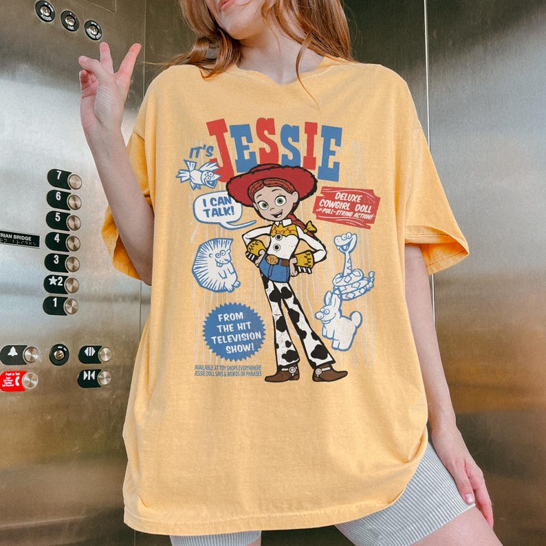 May include: Yellow t-shirt featuring a graphic of the character Jessie from the Toy Story franchise. The design includes the text "Jessie", "I Can Talk!", and "Deluxe Cowgirl Doll". The shirt has short sleeves and a relaxed fit.