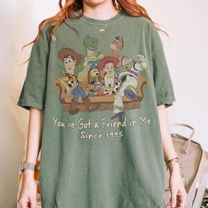 May include: An olive green short-sleeve t-shirt featuring a graphic print of Toy Story characters sitting on a couch. The text reads, "You've Got a Friend in Me" and "Since 1995" in a light-colored font below the illustrated characters.