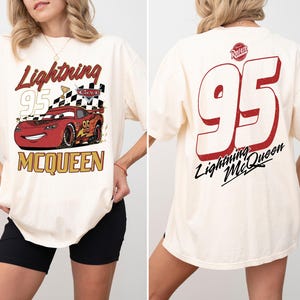 May include: Cream-colored t-shirt featuring a graphic of Lightning McQueen from the Cars movie. The front design includes the text "Lightning 95 McQueen" with a checkered flag. The back displays a large "95" and "Lightning McQueen" text.