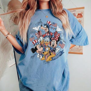 May include: Pale blue t-shirt with a graphic of Disney characters, including Mickey Mouse, Minnie Mouse, Donald Duck, and Pluto, celebrating the 250th USA Anniversary. The design incorporates American flag elements.