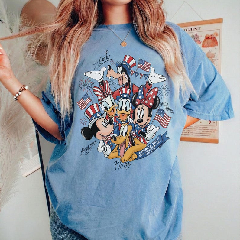 May include: Pale blue t-shirt with a graphic of Disney characters, including Mickey Mouse, Minnie Mouse, Donald Duck, and Pluto, celebrating the 250th USA Anniversary. The design incorporates American flag elements.