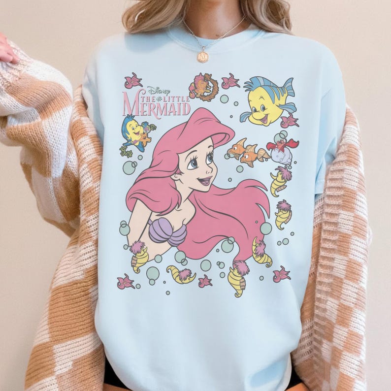 May include: Light blue t-shirt featuring a graphic of Ariel from Disney's The Little Mermaid. Ariel has pink hair and a purple seashell bra. The shirt also includes illustrations of Flounder, Sebastian, and other sea creatures.