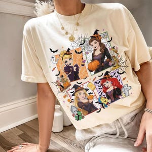 May include: A cream-colored short-sleeve t-shirt featuring a graphic print of four animated characters dressed in Halloween costumes. The design includes spider webs, bats, pumpkins, and tombstones surrounding the characters in colorful frames.