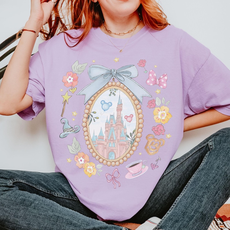 May include: A lilac t-shirt with a fairytale-themed design. The design features an oval frame with a castle, surrounded by floral and bow accents. Additional elements include a teacup, a magic wand, and a bear.