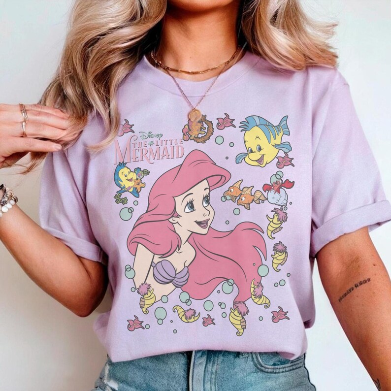 May include: A lilac-colored t-shirt featuring a graphic of Ariel from Disney's The Little Mermaid. The design includes Ariel with pink hair, surrounded by sea creatures like Flounder and Sebastian. The text "Disney The Little Mermaid" is also visible.