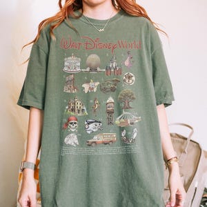 K&ouml;nnte beinhalten: Gr&uuml;nes T-Shirt mit einer Grafik, die verschiedene Symbole der Walt Disney World zeigt, darunter ein Karussell, Spaceship Earth, Cinderella Castle und ein Drache. Die Worte "Walt Disney World" sind rot aufgedruckt.