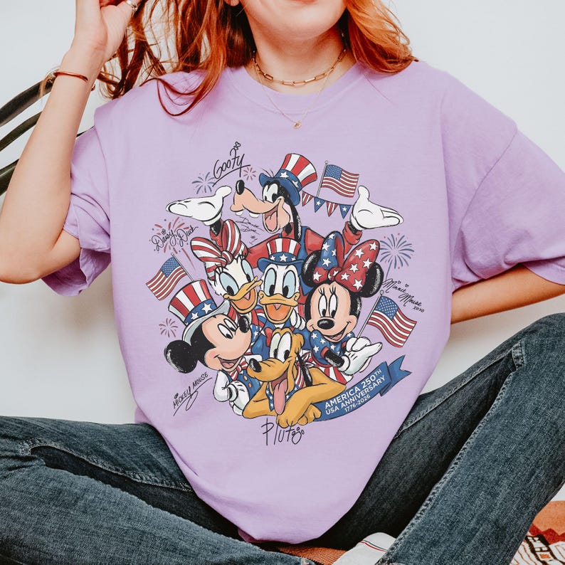 May include: A lilac t-shirt with a graphic of Disney characters Mickey Mouse, Minnie Mouse, Donald Duck, Daisy Duck, Goofy, and Pluto. The design includes American flags and text that reads "America 250th USA Anniversary 1776-2026."