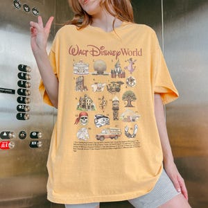 K&ouml;nnte beinhalten: Senfgelbes T-Shirt mit einer Grafik mit verschiedenen Walt Disney World-Symbolen und den Worten "Walt Disney World". Das Shirt hat einen Rundhalsausschnitt und kurze &Auml;rmel.