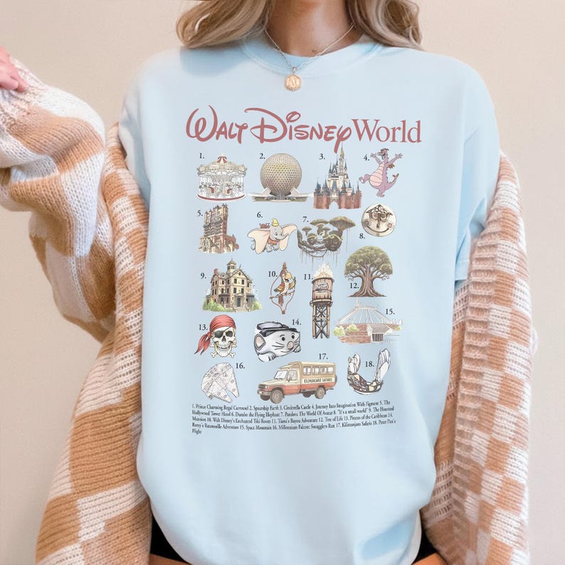 K&ouml;nnte beinhalten: Hellblaues T-Shirt mit einer Grafik von Walt Disney World-Attraktionen. Das Design umfasst Illustrationen von Cinderella's Castle, Spaceship Earth und anderen ikonischen Wahrzeichen. Der Text "Walt Disney World" ist oben in Burgunderrot gedruckt.