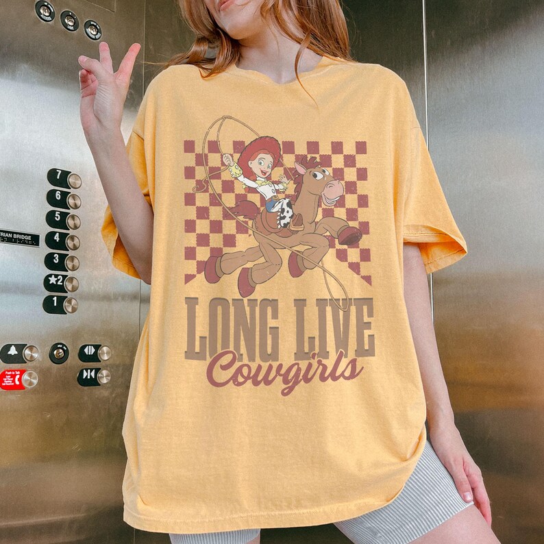 K&ouml;nnte beinhalten: Senfgelbes T-Shirt mit einem Cartoon-Cowgirl, das mit einem Lasso auf einem Pferd reitet, vor einem karierten Hintergrund. Der Text "LONG LIVE Cowgirls" ist darunter gedruckt. Das T-Shirt hat kurze &Auml;rmel.