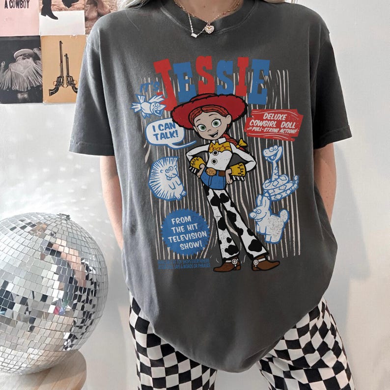 May include: Dark gray t-shirt featuring a graphic of the character Jessie from the Toy Story franchise. The design includes the text "Jessie", "I Can Talk!", and "Deluxe Cowgirl Doll". The shirt also has the text "From the Hit Television Show!"