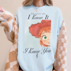 K&ouml;nnte beinhalten: Ein hellblaues Sweatshirt mit Rundhalsausschnitt und einer Grafik einer rothaarigen Figur mit rotem Cowboyhut. Der Text lautet "EST 2026 I Knew It I Knew You" mit einem Violinschl&uuml;ssel unter dem Text. Das Sweatshirt wird mit einer beigen Strickjacke getragen.