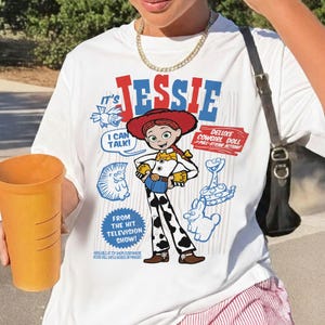 May include: White t-shirt featuring a graphic of Jessie from the "Toy Story" franchise. The design includes the text "It's Jessie," "I Can Talk!" and "Deluxe Cowgirl Doll." Additional blue cartoon graphics and text are also present.