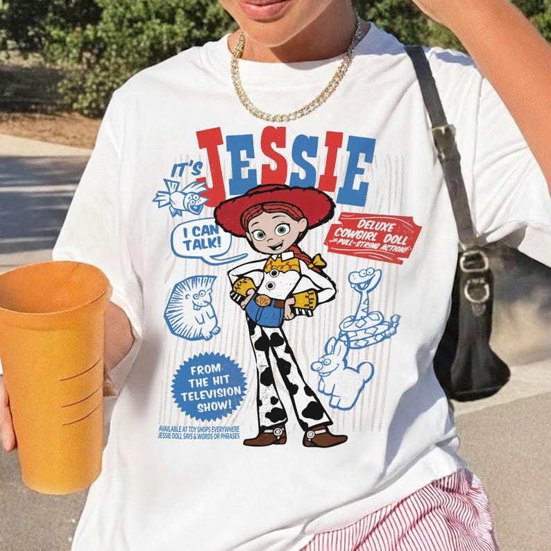 May include: White t-shirt featuring a graphic of Jessie from the "Toy Story" franchise. The design includes the text "It's Jessie," "I Can Talk!" and "Deluxe Cowgirl Doll." Additional blue cartoon graphics and text are also present.