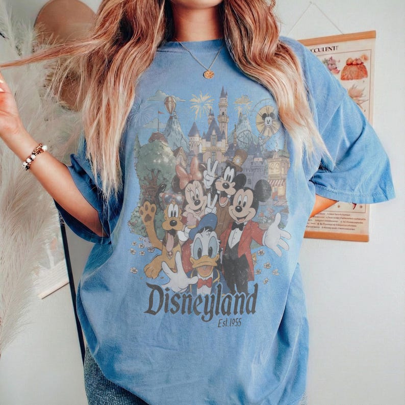 Puede incluir: Camiseta azul claro con un gr&aacute;fico de Disneyland con Mickey Mouse, Minnie Mouse, Donald Duck y Goofy. El gr&aacute;fico incluye el logotipo de Disneyland y "Est. 1955". La camiseta tiene mangas cortas y un corte holgado.