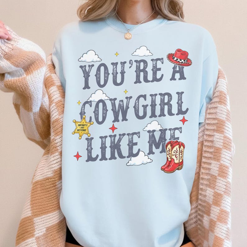 May include: A light blue sweatshirt featuring the text "YOU'RE A COWGIRL LIKE ME" in a dark gray font. The design includes white clouds, small yellow and red stars, a yellow sheriff badge with text, a red cowboy hat, and red cowboy boots.