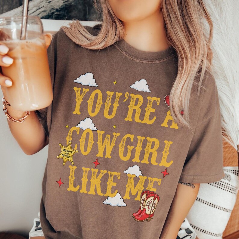 May include: A brown short-sleeve t-shirt featuring the yellow text "YOU'RE A COWGIRL LIKE ME" with white cloud graphics, small red stars, a yellow sheriff star badge, and a pair of red and white cowboy boots printed on the front chest area.