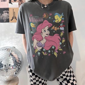 May include: Dark gray t-shirt featuring Ariel from Disney's The Little Mermaid. The graphic includes Ariel with pink hair, a purple seashell bra, and various sea creatures. The shirt has the movie title printed above the character.